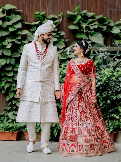 A beautiful walking portrait of the couple in their engagement attire. The bride's red lehenga and the groom's white sherwani create a classic and elegant look against the green backdrop.
