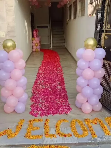A beautiful and traditional welcome entrance for a baby. The path is decorated with a carpet of fresh flower petals, flanked by pink and lilac balloon pillars, and a 'WELCOME' message written with marigold flowers.