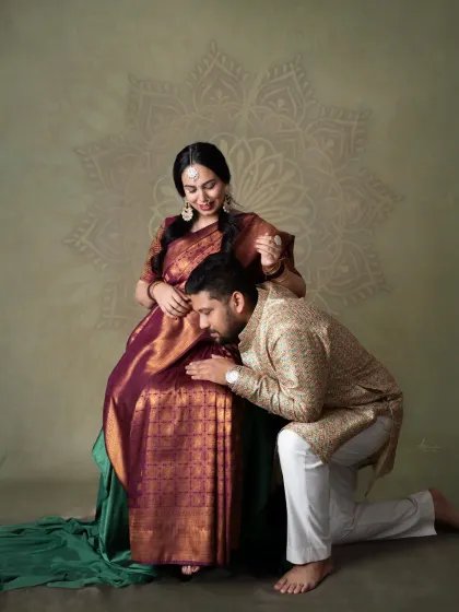A tender gesture of love and respect. The dad-to-be kneels to kiss the baby bump, a beautiful moment captured in their traditional attire.