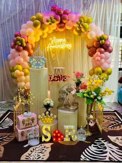 A colorful anniversary setup with a mix of warm-toned balloons, a neon sign, and various props like a mini Ferris wheel and bird cages for a whimsical touch.