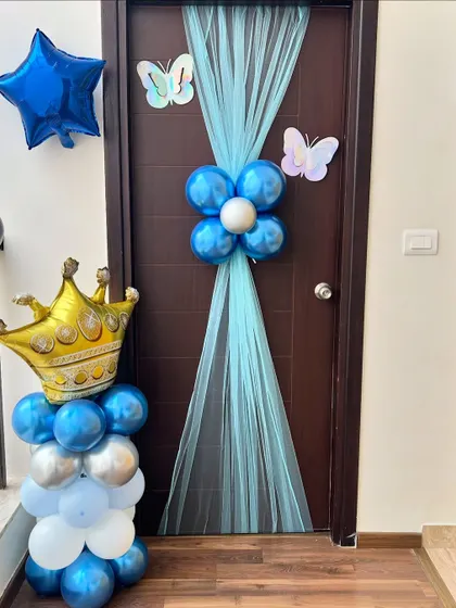 A simple and sweet door decoration to welcome a new baby boy home. The arrangement includes a blue and silver balloon column topped with a gold crown and elegant blue tulle.