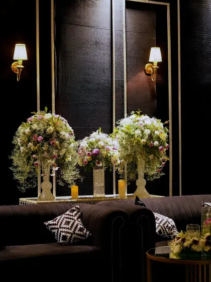 A chic lounge setting with plush black sofas and large white floral arrangements. The gold-trimmed wall panels and classic sconces add a touch of art deco glamour.