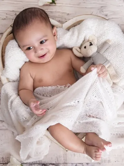 An adorable baby boy, a few months old, holding a tiny teddy bear while lying in a heart-shaped basket.