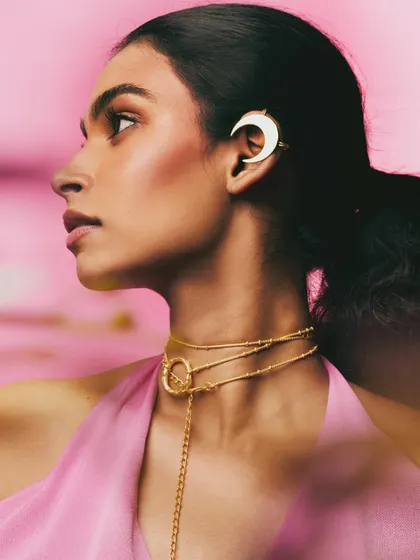 A profile shot for a Batoki.art jewelry campaign, set against a soft pink background. The focus is on the delicate gold necklace and a unique crescent moon ear cuff, highlighting the brand's artistic designs.
