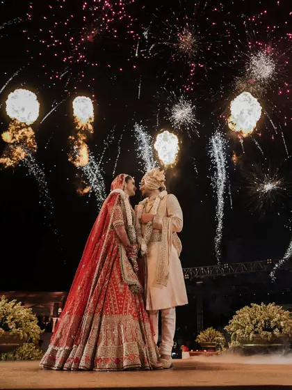 A fairytale moment under the fireworks. The bride's magnificent red wedding lehenga and the groom's classic sherwani create a picture-perfect memory against the night sky.