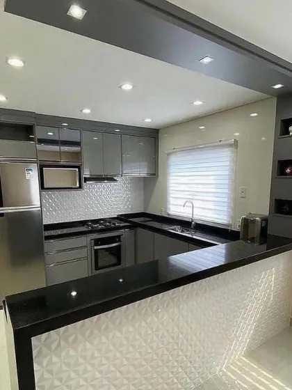 An open-plan kitchen with a U-shaped layout and an attached breakfast bar. The combination of glossy grey cabinets, a black granite countertop, and a 3D textured white backsplash creates a bold, modern statement.