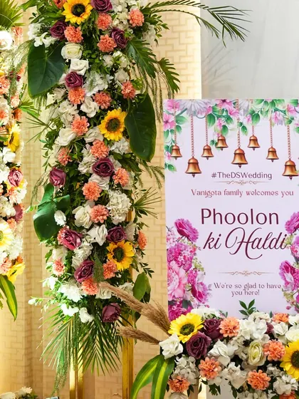 The welcome signage for the 'Phoolon ki Haldi', featuring bright floral graphics and traditional temple bells. This set a festive and welcoming tone for the event right from the entrance.