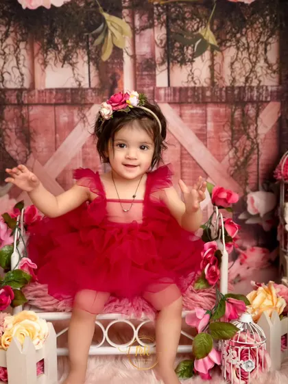 A toddler girl in a festive red dress sits on a miniature bed surrounded by flowers for her birthday shoot.