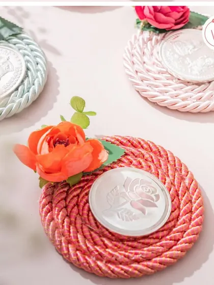 A creative and beautiful way to present a silver coin favor, mounted on a colorful, braided coaster and adorned with a flower.