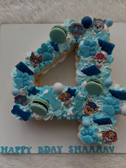 A Paw Patrol-themed number cake for a 4th birthday. The '4' is decorated with blue frosting, macarons, and all the favorite pup characters.