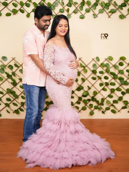 A lovely couple's portrait in our studio. The mom-to-be wears a beautiful pink lace and tulle gown, standing with her partner against a green vine wall for a touch of nature indoors.