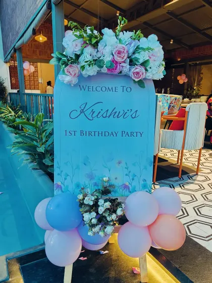 A custom-shaped welcome board for a first birthday party. The sign is topped with a beautiful artificial flower arrangement and surrounded by balloons.
