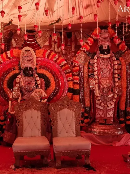 A divine wedding stage featuring large idols of Lord Venkateswara and Goddess Padmavathi. The backdrop is richly decorated with concentric circles of flowers and traditional garlands, creating a sacred ambiance.