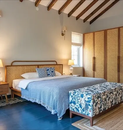 This bedroom in our Goa villa project embraces rustic charm with a modern touch. The custom bed features a natural rattan headboard, paired with oak side tables and a vibrant floral bench. The exposed wooden ceiling beams add to the room's character.