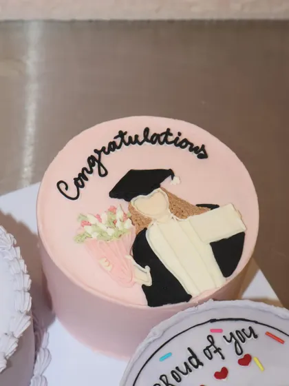 This personalized graduation bento cake features a hand-piped illustration of the graduate in her cap and gown, holding a bouquet of flowers. It's a sweet and personal way to say "Congratulations!".