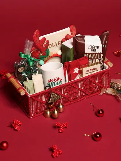 This cozy Christmas hamper is packed in a red macrame basket and includes a 'Sleigh All Day' mug, waffle chips, candles, and other festive treats.