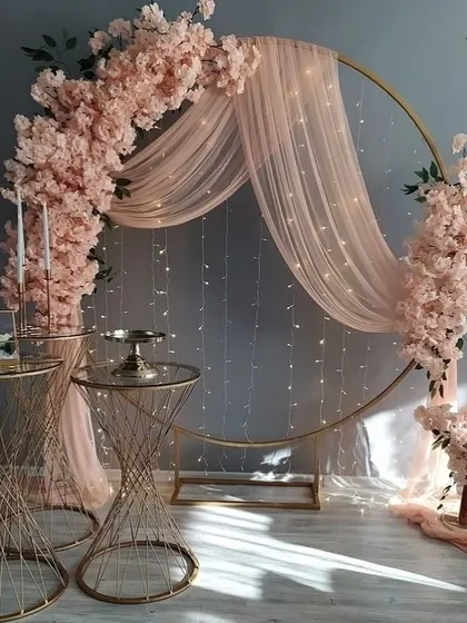 An inspirational design for a romantic photo booth, featuring a large golden ring adorned with pink cherry blossoms, fairy lights, and sheer drapes.