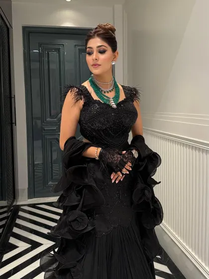 The structured updo adds height and elegance, balancing the voluminous ruffles of the gown and drawing attention to the emerald statement necklace.