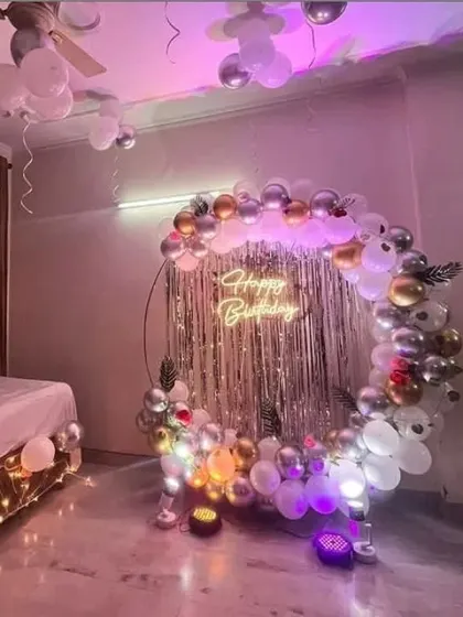 A birthday party in a bedroom with a circular balloon arch in silver, gold, and white. The setup includes a silver fringe backdrop and a "Happy Birthday" neon sign.