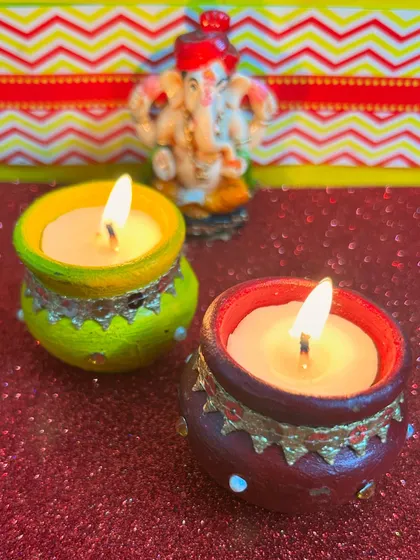 A pair of lit matki candles in vibrant colors, with a mini Ganesha in the background. Perfect for your Diwali pooja.