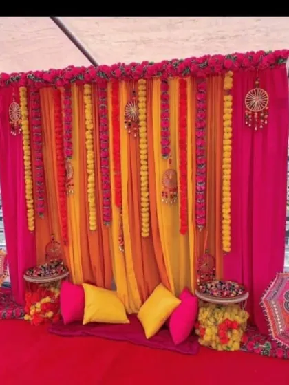 A cozy Mehendi seating area with a backdrop of pink and yellow drapes and hanging marigold garlands. The setup includes floor seating with colorful cushions and traditional embroidered umbrellas.