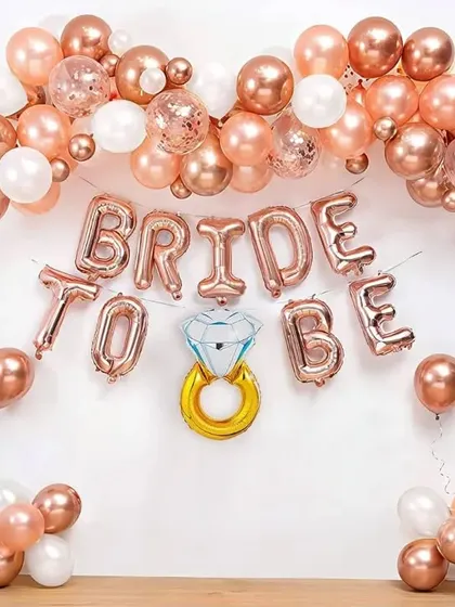 A DIY-style "BRIDE TO BE" decoration against a simple white wall. This setup uses rose gold foil letter balloons, a diamond ring foil balloon, and a matching balloon garland, perfect for a simple and chic celebration at home.