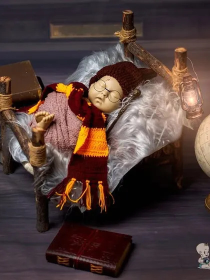 A magical newborn photoshoot with a Harry Potter theme. The baby is sleeping peacefully on a tiny rustic bed, wearing a Gryffindor scarf, next to a globe and a stack of old books.