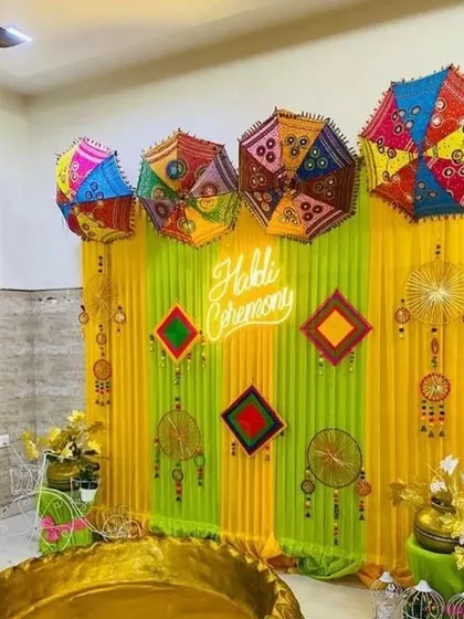 An indoor Haldi ceremony corner with a large golden 'urli' and a backdrop of green and yellow drapes, decorated with umbrellas and a neon "Haldi Ceremony" sign.