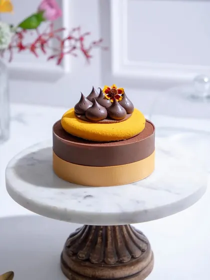 A sophisticated two-toned entremet in shades of brown and yellow, topped with chocolate quenelles and a marigold flower.