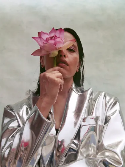 A conceptual portrait where a metallic silver jacket is paired with a natural element, a pink lotus. The styling creates a beautiful contrast between the hard, reflective surface of the jacket and the soft, organic texture of the flower.