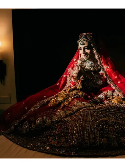A dramatic, low-angle seated portrait of the bride. The lighting highlights the vastness and intricate detail of her red bridal lehenga.