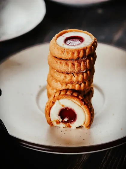 A close-up shot of jam-filled cookies stacked in a tower. The moody lighting and tight focus on the bite taken out of the front cookie create a sense of indulgence and rustic charm.