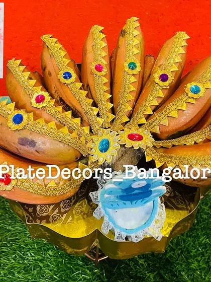 A bunch of bananas decorated with golden fabric and colorful kundan stones, with a small blue cradle cutout for a baby boy ceremony.