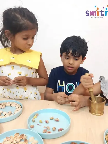 A boy carefully uses a mortar and pestle, a practical life skill that builds strength and coordination. His friend watches intently, learning through observation.