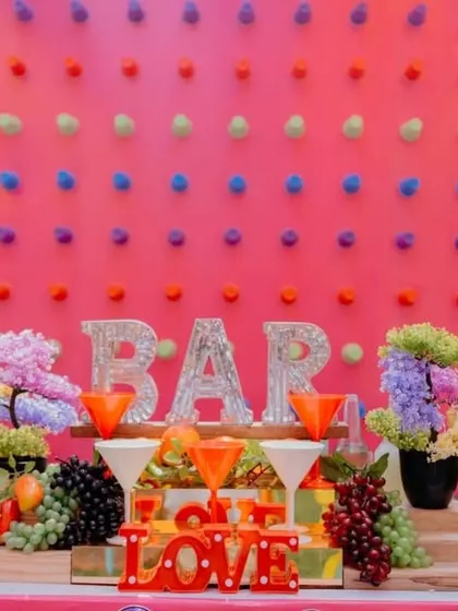 A detailed view of the bar counter. The setup includes fun props like light-up "BAR" and "LOVE" signs, wooden barrels, and fresh fruit, adding to the celebratory vibe.