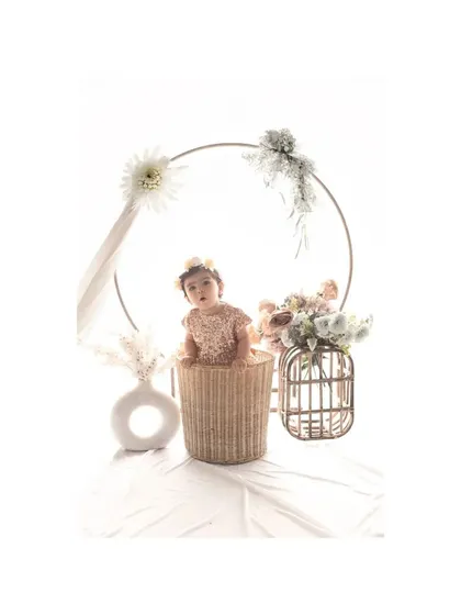 A baby girl sits in a wicker basket, framed by a large golden ring adorned with flowers. This elegant and artistic studio setup creates a beautiful and memorable portrait for a special milestone.