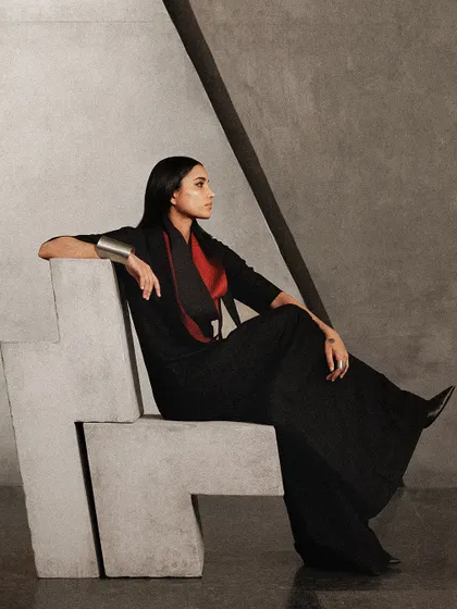 The model is seated on a custom-designed concrete chair, blurring the line between furniture, sculpture, and fashion. The styling is strong and statuesque.