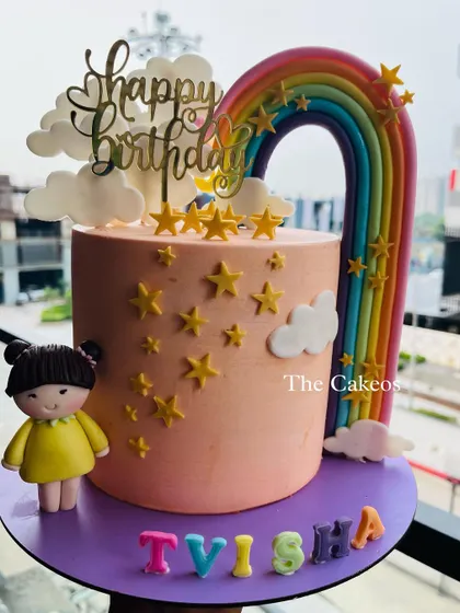 A tall and elegant cake with a rainbow and doll topper. The peach-colored cake is decorated with stars and clouds for a dreamy birthday theme.