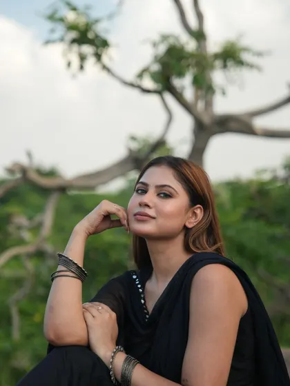 A simple and natural outdoor portrait of a model in a black suit, sitting on a rock with greenery in the background.