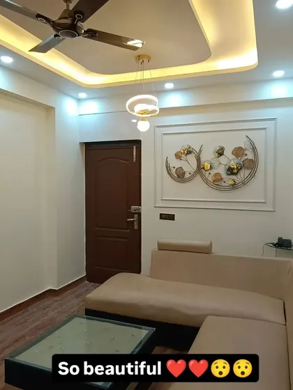 The seating area of the same Noida Sector 62 project, showing the L-shaped sofa, simple false ceiling with modern lighting, and a beautiful metal wall art piece.