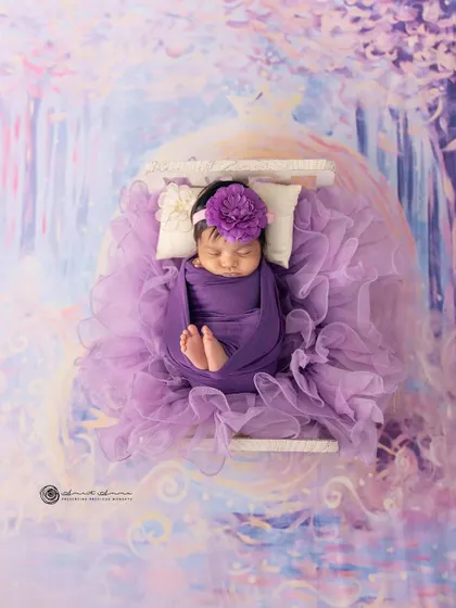 A lavender dream. This newborn is nestled in a bed of purple tulle against a magical, painted backdrop, creating a truly enchanting and artistic portrait.