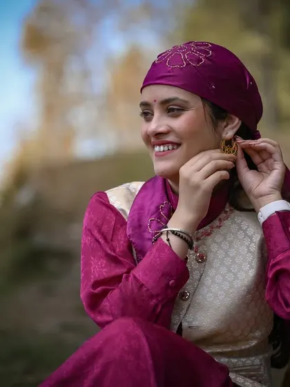 A close-up portrait of the woman in her vibrant traditional dress, smiling as she adjusts her earring. This shot highlights the intricate details of Pahari jewelry and attire.