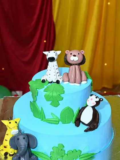A delightful two-tier jungle-themed cake. It features cute, edible animal figures like a zebra, lion, and monkey, perfectly matching the party's theme.