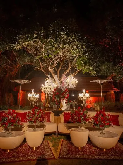 A beautiful symmetrical lounge setup under a large tree. The curved white sofas are arranged on a traditional red carpet, with candelabras and floral arrangements creating a regal, darbar-like feel.
