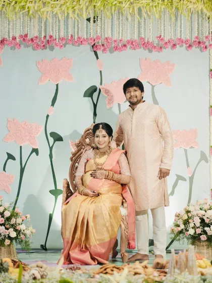 A modern take on a traditional baby shower, with a beautiful lotus-themed backdrop in soft pastel colors. The mother-to-be is seated on an elegant, traditional chair.