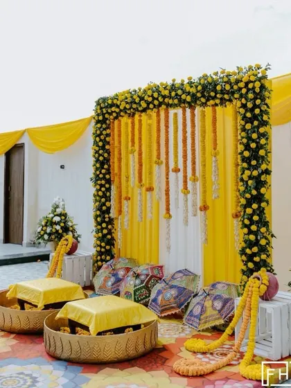 A beautiful and simple Haldi seating area with low stools placed in decorative baskets. The backdrop is a classic frame of marigolds with hanging floral strings, perfect for an intimate home ceremony.