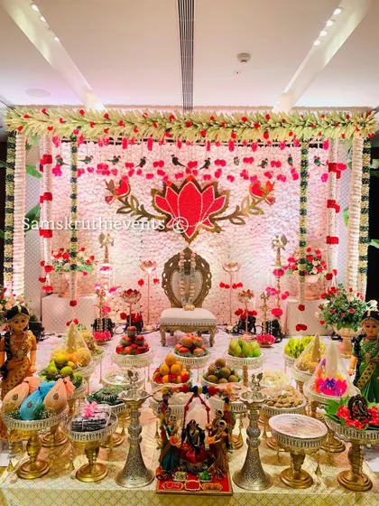 A grand and festive setup for a half-saree or puberty ceremony. The backdrop features a large lotus motif made of flowers, surrounded by traditional brass lamps and a beautiful display of offerings, marking the milestone in style.