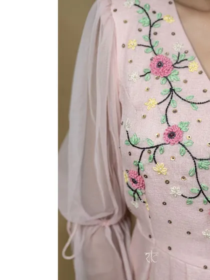 A close-up of a blush pink floor-length dress, showcasing the delicate floral thread embroidery on the bodice. The sheer puff sleeves add a touch of romantic charm.