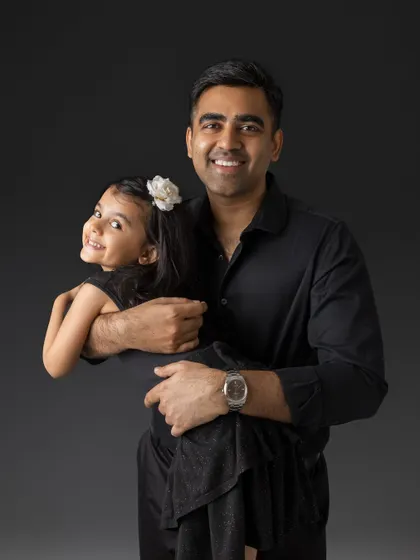 A joyful portrait of a father and his daughter. He holds her in his arms as she peeks over his shoulder with a big smile, a happy moment captured during their family maternity session.