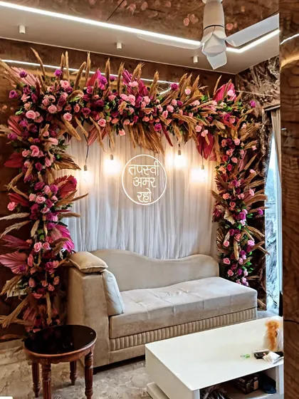 A beautiful floral arch in shades of pink and magenta, designed for a Tapasvi ceremony in a living room. The warm lighting and dried palm leaves create a sophisticated and modern look.